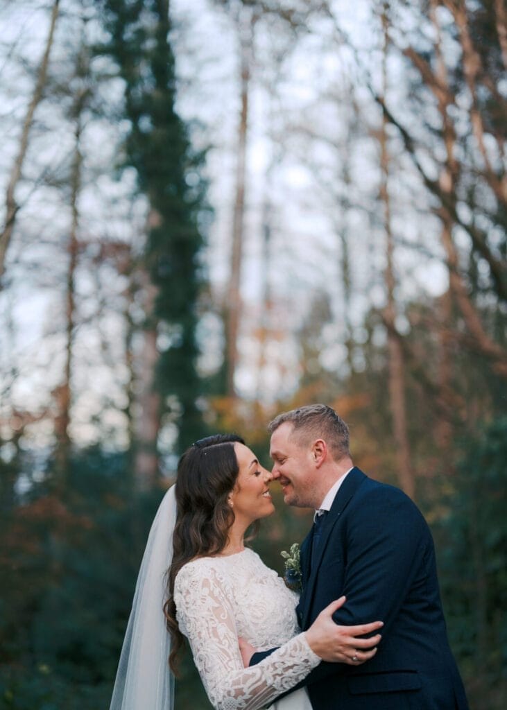 In the heart of Thunderbridge, a couple dressed in wedding attire shares an intimate moment. The bride, in a lace gown and veil, and the groom, in a dark suit, are embraced forehead to forehead among towering trees—a perfect capture for The Woodman Inn wedding photographer.