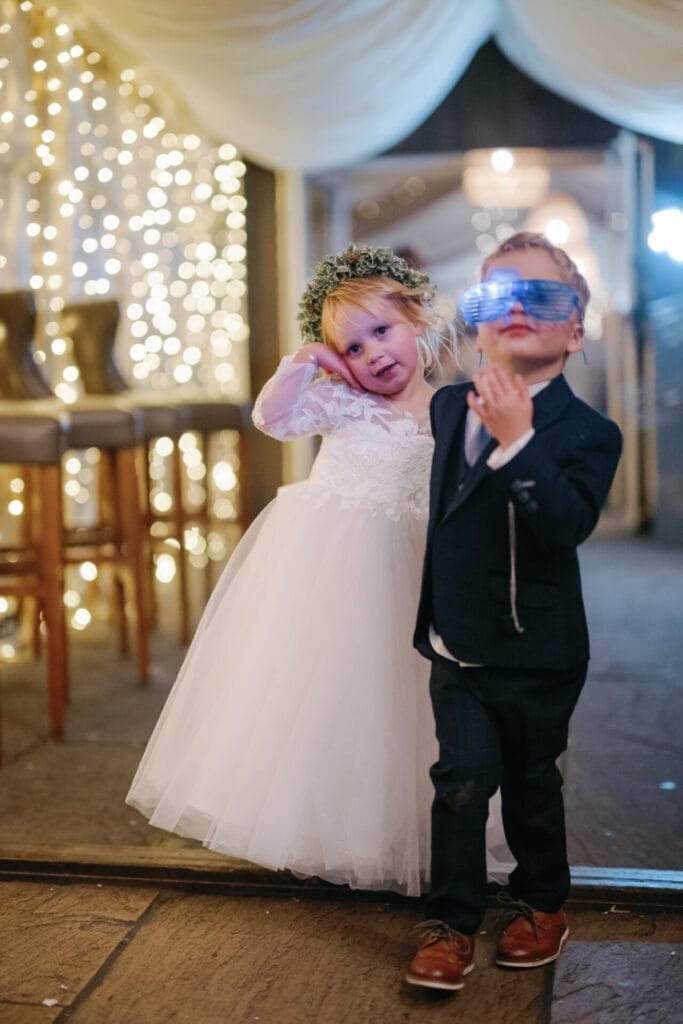 At a charming Thunderbridge wedding, two children steal the spotlight. A girl in a white dress and floral headpiece smiles sweetly, hand on cheek. Next to her, a boy in a suit and blue sunglasses exudes confidence. The warm fairy lights, captured perfectly by The Woodman Inn wedding photographer, twinkle behind them.