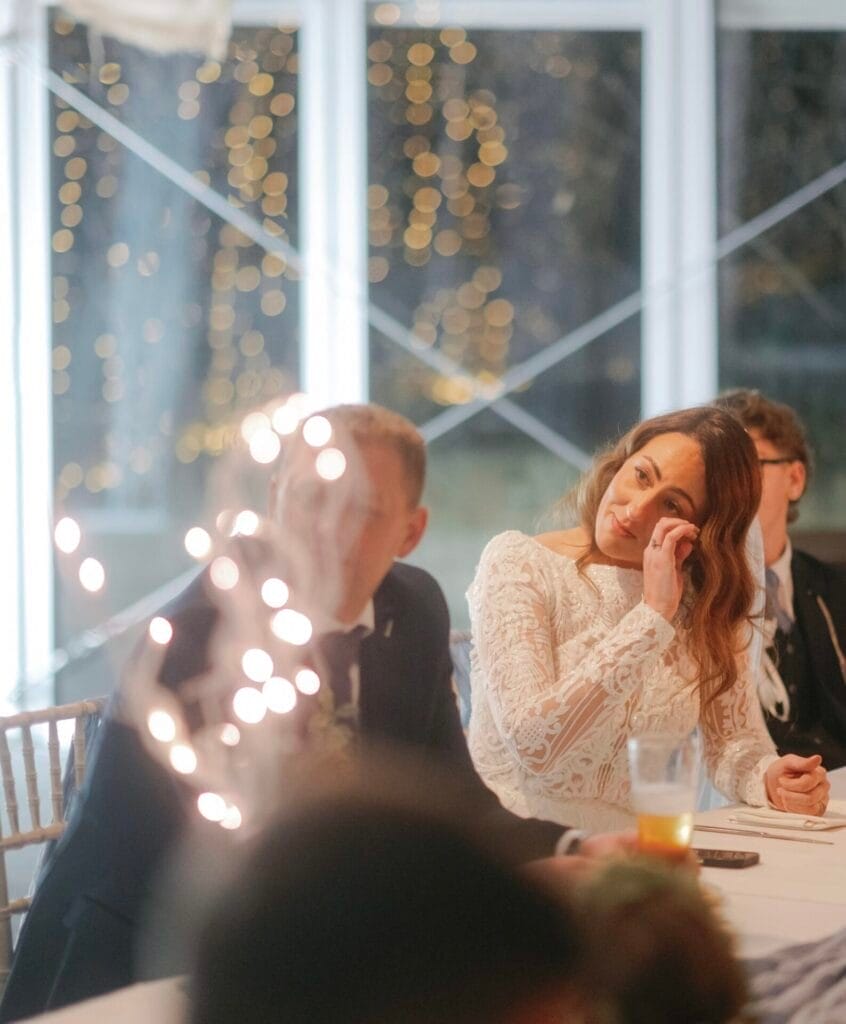 A woman in a white lace dress sits at a table, resting her head on her hand, listening attentively beside a man in a suit. The Woodman Inn wedding photographer captures the softly lit backdrop with blurred lights, adding warmth and intimacy to the Thunderbridge setting.