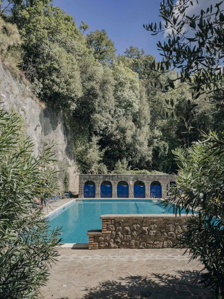 A serene outdoor swimming pool at Villa Palazzola is surrounded by lush greenery and stone walls. Arched doorways line the far side, reflecting in the clear blue water. Shaded by trees, this tranquil Italian destination near Rome offers a secluded haven for any discerning wedding photographer.