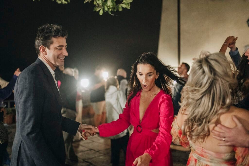 At a Villa Palazzola wedding near Rome, a man in a suit and a woman in a red dress dance energetically at an outdoor evening event. The background is filled with people mingling and dancing under soft lights, creating a festive atmosphere perfect for any photographer in Italy to capture.