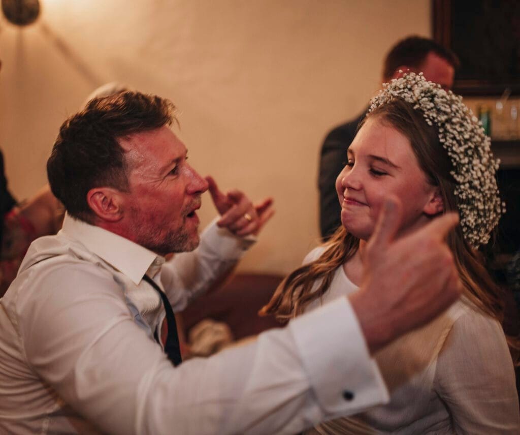 At a vibrant Villa Palazzola, a man in a white shirt joyfully interacts with a young girl wearing a floral crown. The festive setting hints at a special event, possibly captured by a talented wedding photographer in Italy. Both are smiling and engaging with each other warmly.