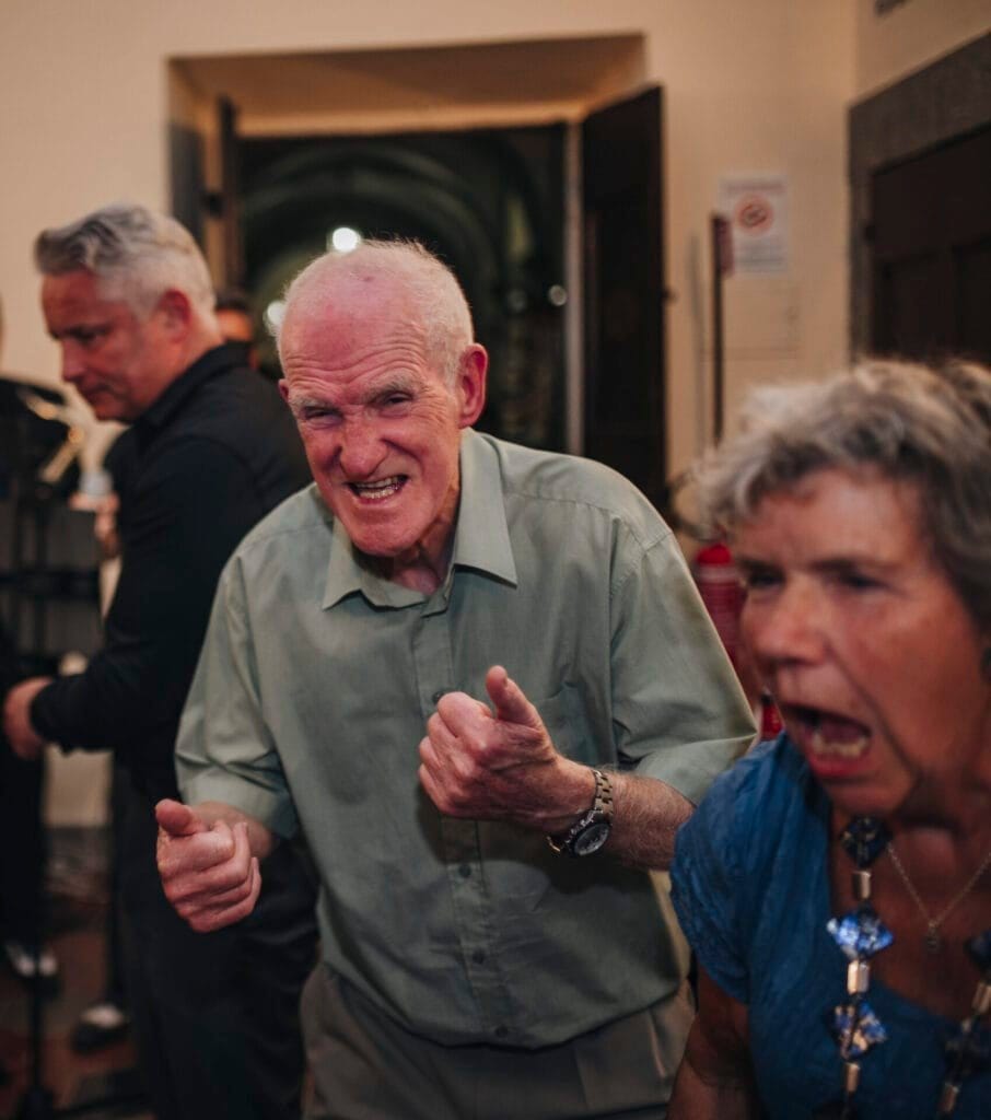 An older man in a green shirt, embodying playful intensity with fists up, stands beside a woman in a blue top mirroring his lively expression. In the dim indoor setting of Villa Palazzola near Rome, another man in black lingers behind, captured by a wedding photographer in Italy.