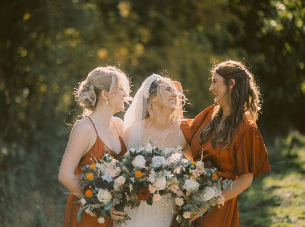Three smiling women pose outdoors in warm lighting, surrounded by greenery. The bride, in the center, wears a white dress and veil, holding a bouquet. The two bridesmaids wear matching rust-colored dresses, each holding a similar bouquet.