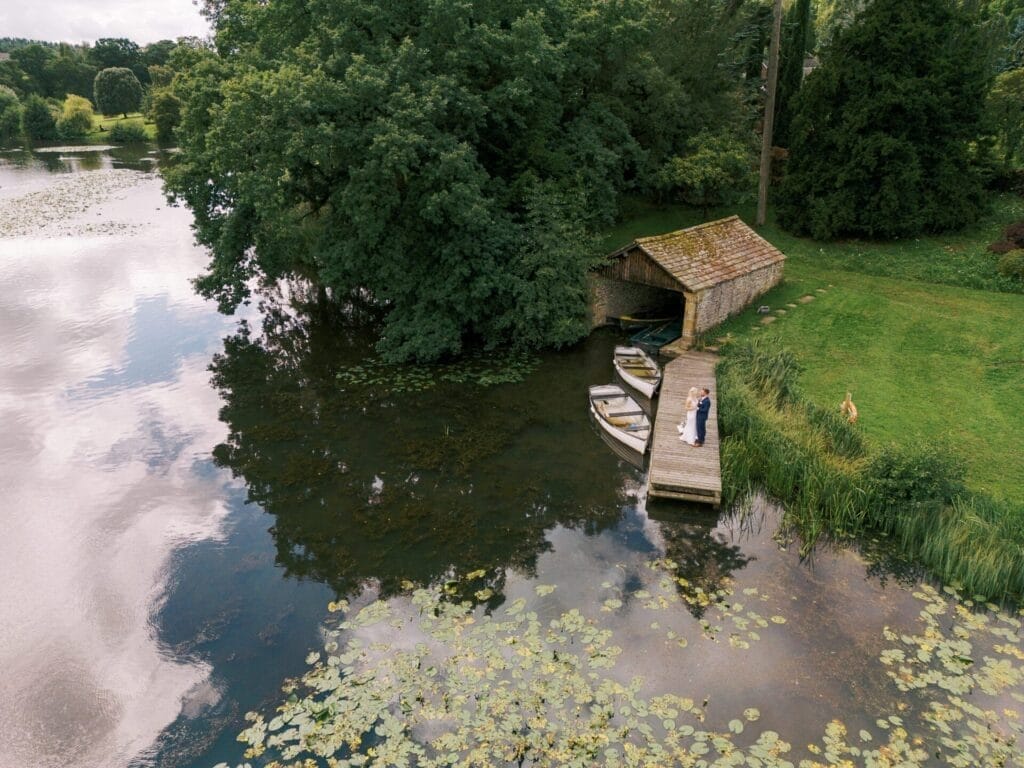 Aerial view of a couple on a wooden dock by a small, rustic boathouse. Three rowboats are moored beside them. The scene is surrounded by lush greenery and a calm pond with lily pads, reflecting the cloudy sky above.