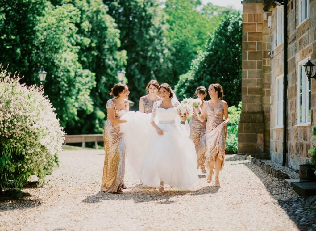 A bride in a white gown walks joyfully with four bridesmaids in shimmering dresses. They are outside on a sunny day, surrounded by greenery and a stone building. The bridesmaids help carry the brides train as they walk on a gravel path.