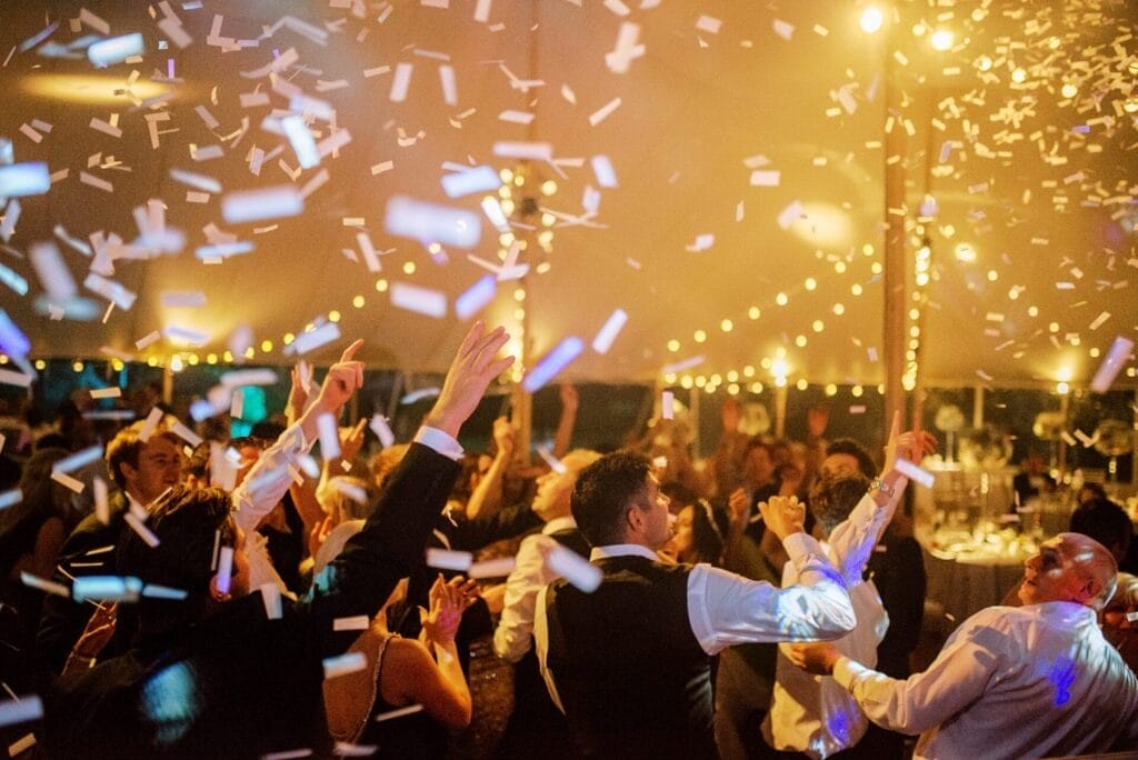 People are celebrating at a festive indoor party with confetti falling from above. Some are raising their hands, while others are dancing. Warm lights decorate the ceiling, creating a joyful and lively atmosphere.