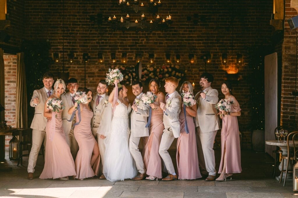 A joyful wedding party poses in an indoor setting. The bride and groom are in the center, surrounded by bridesmaids in pink dresses and groomsmen in beige suits. Everyone is laughing and holding bouquets. The background features brick walls and hanging lights.