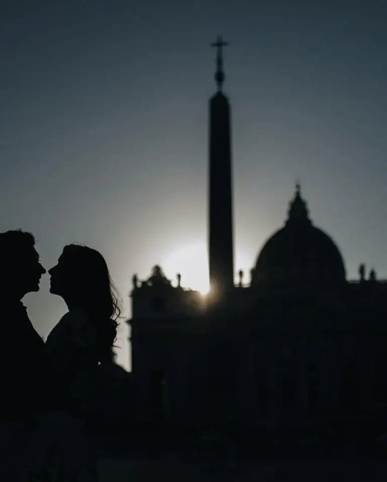 Silhouette of two people facing each other in front of St. Peters Basilica at sunset, with the sun setting behind the dome and obelisk.