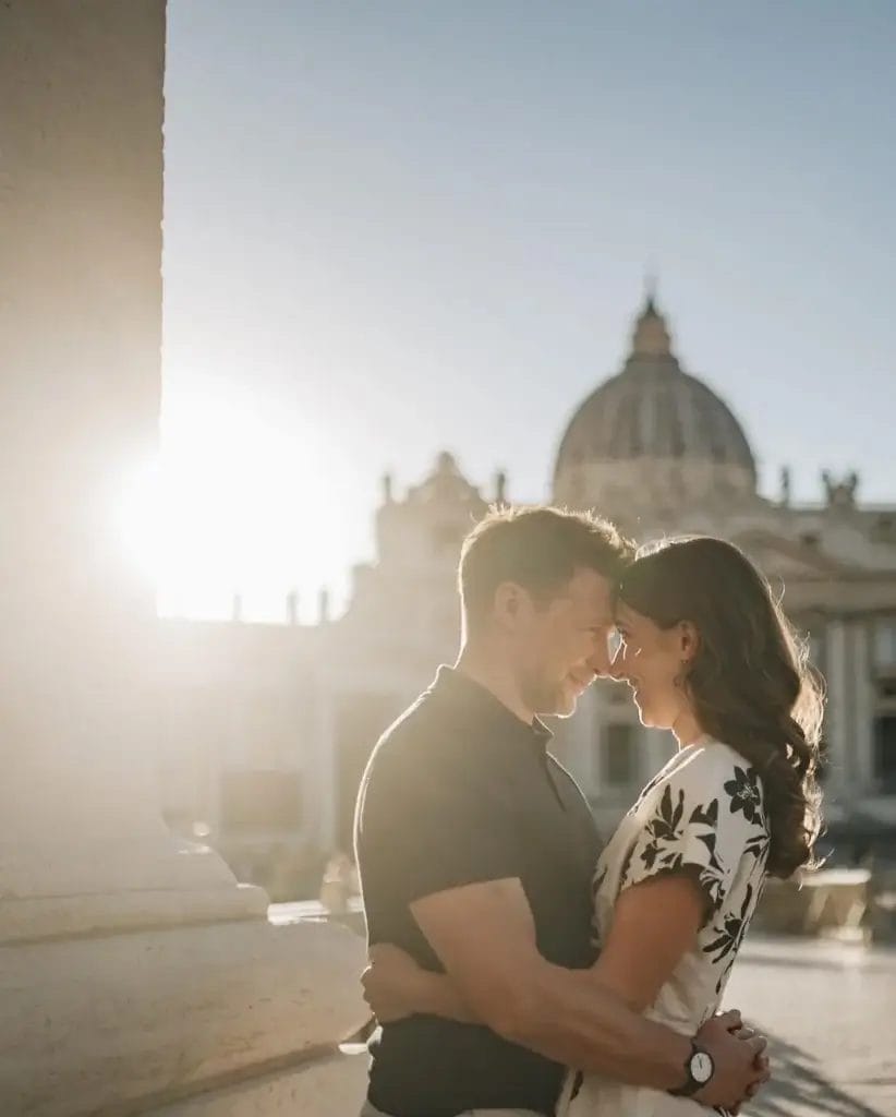 Yorkshire Engagement Photographer 10 A couple stands close, embracing and smiling, with their foreheads touching. They are outdoors with a blurred, historic building and clear sky in the background. The sunlight creates a soft glow around them.