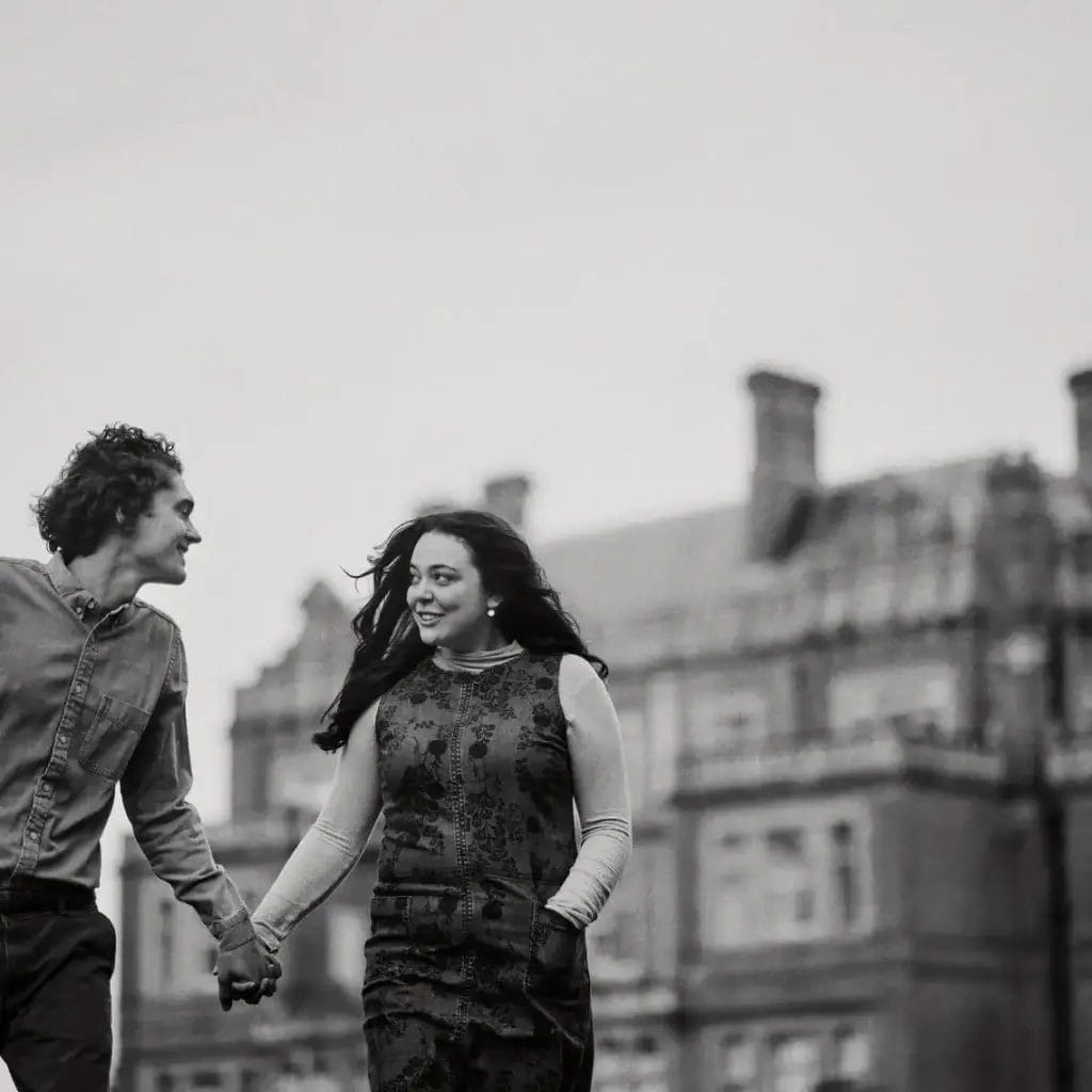 Yorkshire Engagement Photographer 12 A black and white photo showing a smiling couple holding hands and walking. The man is wearing a shirt and pants, while the woman is in a long dress. They are in front of a large, historic building with a cloudy sky above.