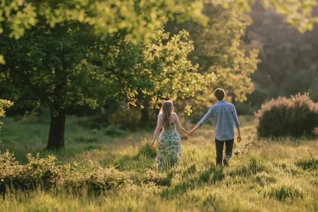 Yorkshire Engagement Photographer 9 A couple holding hands walks through a sunlit grassy field. The woman wears a floral dress, and the man is in a blue shirt. They are surrounded by trees and golden light filtering through the leaves.