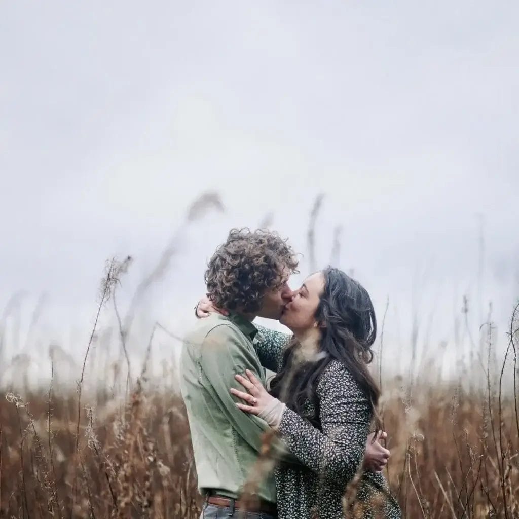 Yorkshire Engagement Photographer 14 A couple kisses in a field of tall, dry grasses under a cloudy sky. The man has curly hair and wears a green shirt, while the woman has long dark hair and wears a patterned coat.