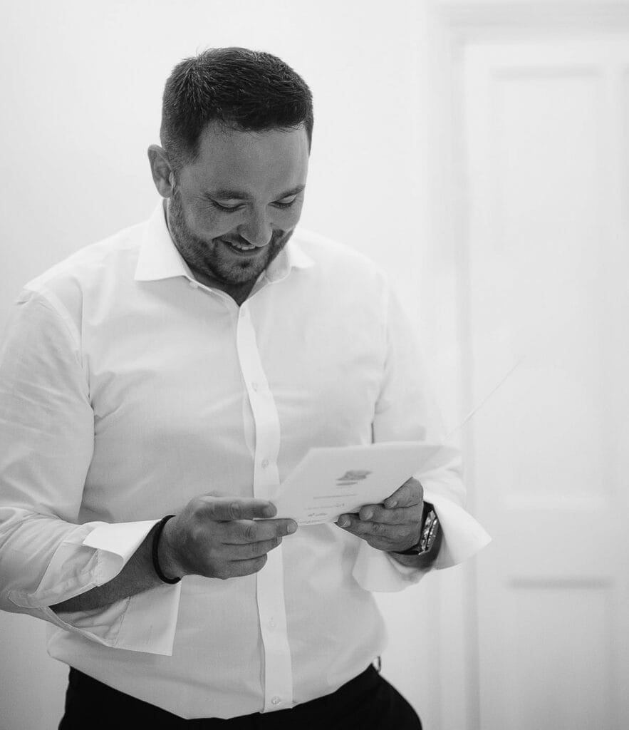 A man in a white shirt, smiling as he reads a card, stands near a bright wall and closed door, appearing happy and engaged with the cards contents. This charming black-and-white moment captures the essence of joy found in Hornington Manor wedding photos.