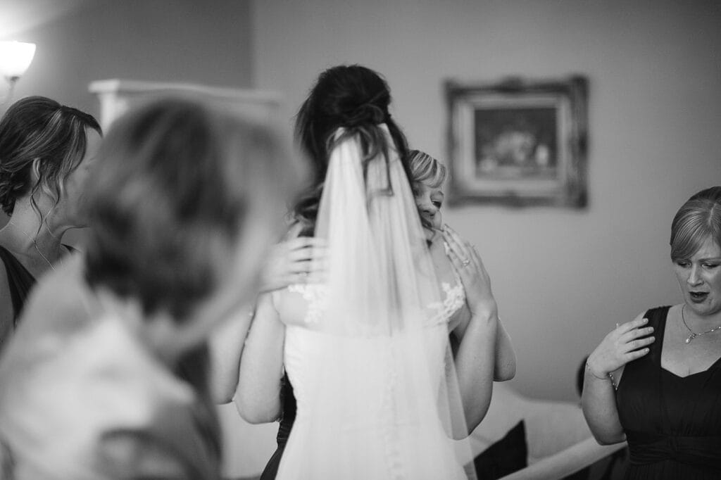 In the warmly lit room, a bride in a wedding gown and veil is lovingly embraced by another woman. Surrounded by smiling women, one appears emotional. Captured by a Hornington Manor wedding photographer, a framed picture hangs elegantly on the wall in the background.