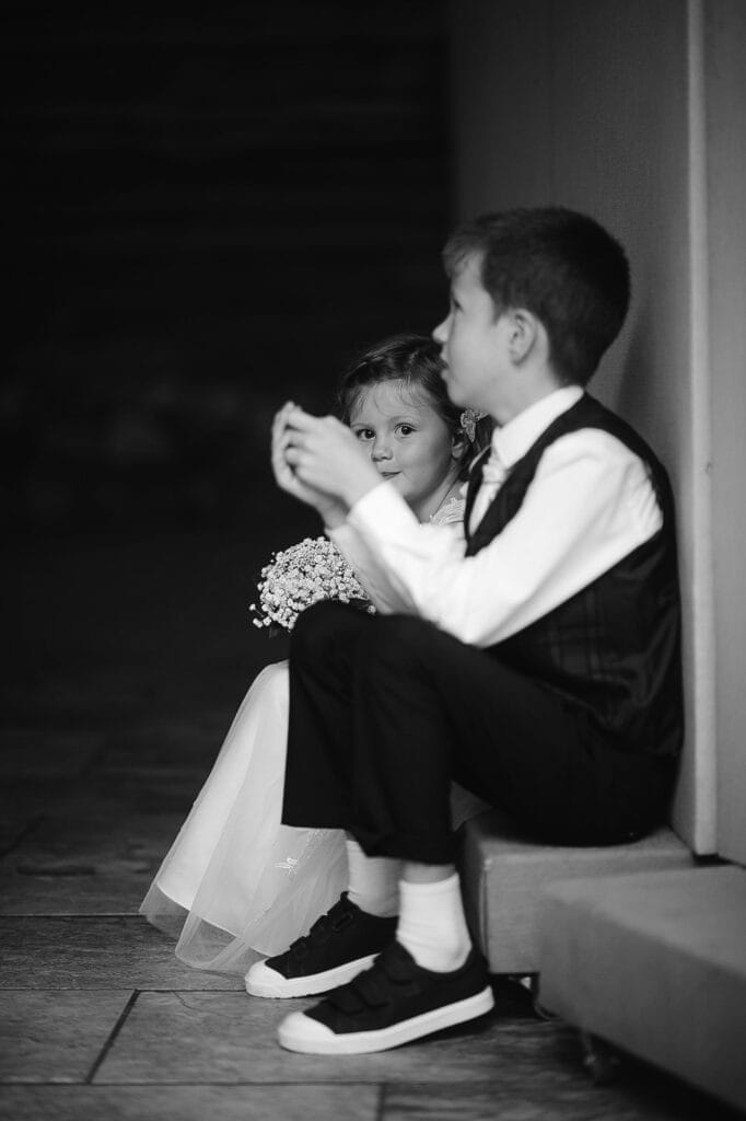 A black and white photo captures a young boy and girl at Hornington Manor. The girl, wearing a dress and holding flowers, gazes thoughtfully ahead. Beside her, the boy in formal attire looks away, both seated on an elegant stone surface—a timeless moment skillfully framed by the wedding photographer.
