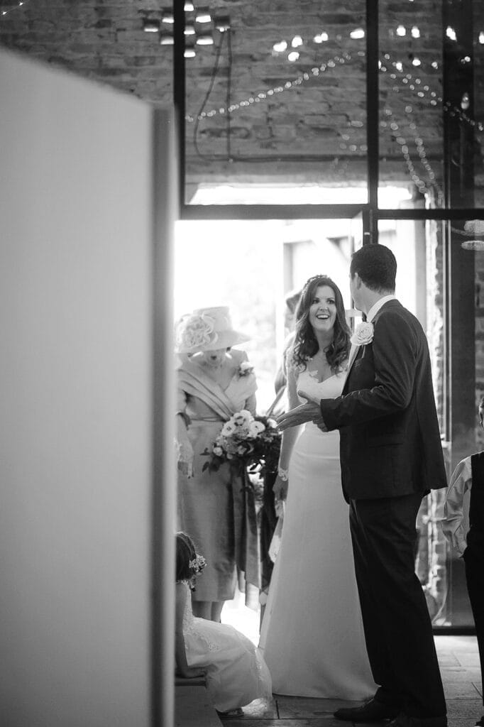 A bride and groom stand smiling inside a rustic venue with large glass doors. The bride holds a bouquet, and an older woman wearing a hat stands nearby. Captured by a Hornington Manor wedding photographer, the black-and-white photo reflects joy with soft natural light.