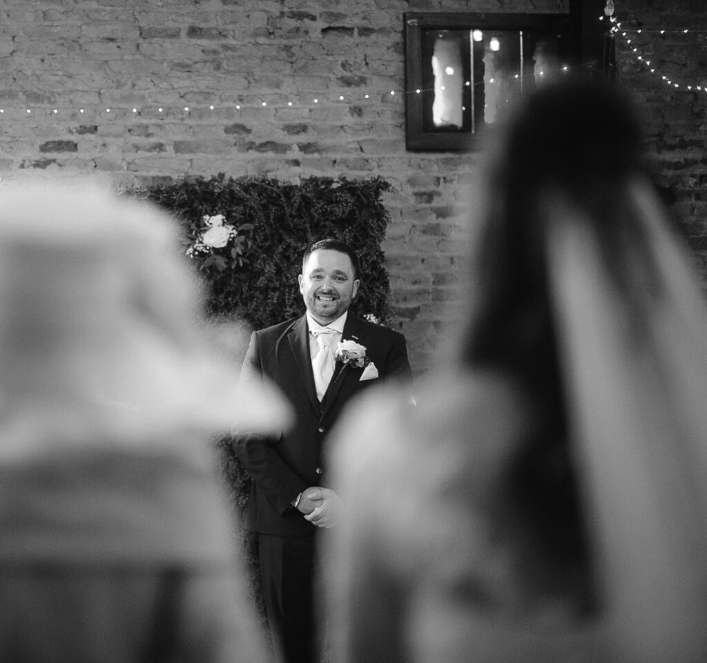 Groom in a suit and boutonniere stands smiling in the rustic brick-walled venue at Hornington Manor, with blurred figures in the foreground. Festive lights and floral decorations enhance the warm ambiance, perfectly captured by the wedding photographer.