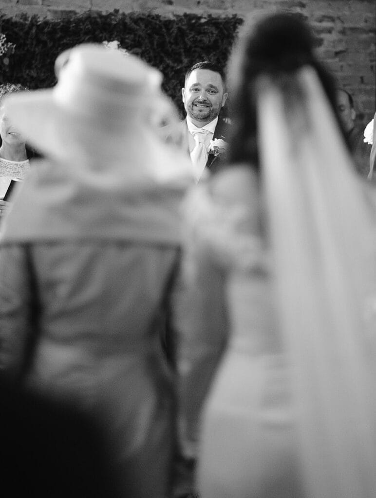 Black and white photo of a Hornington Manor wedding ceremony. The groom is smiling at the bride, who is wearing a veil. A person in a hat stands nearby. The focus is on the grooms expression, with the lush greenery enhancing this captured moment.