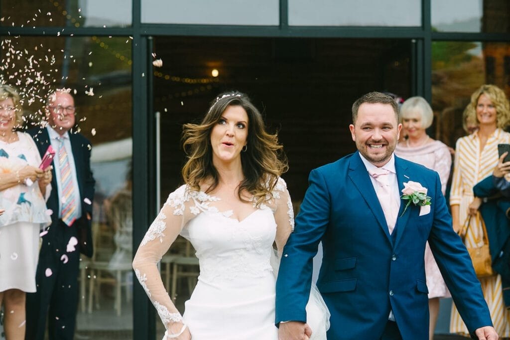 A joyful bride and groom walk hand in hand, smiling, as wedding guests shower them with confetti. Captured by the Hornington Manor wedding photographer, the bride dazzles in a white dress with lace sleeves, while the groom stuns in a navy suit. Guests smile and clap warmly in the background.