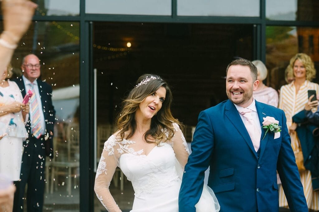 A bride and groom are smiling joyfully as they walk past onlookers at Hornington Manor. The bride wears a white lace dress, and the groom sports a navy suit with a pink tie and boutonniere. Confetti flutters through the air while people clap in the background, captured by their wedding photographer.