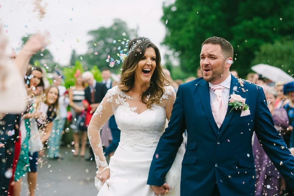 Bride and groom walking outdoors, surrounded by guests showering them with confetti. The bride dazzles in a white gown with lace sleeves, and the groom stands out in a blue suit with a pink tie. Both are all smiles against the green, lively backdrop captured by the Hornington Manor wedding photographer.
