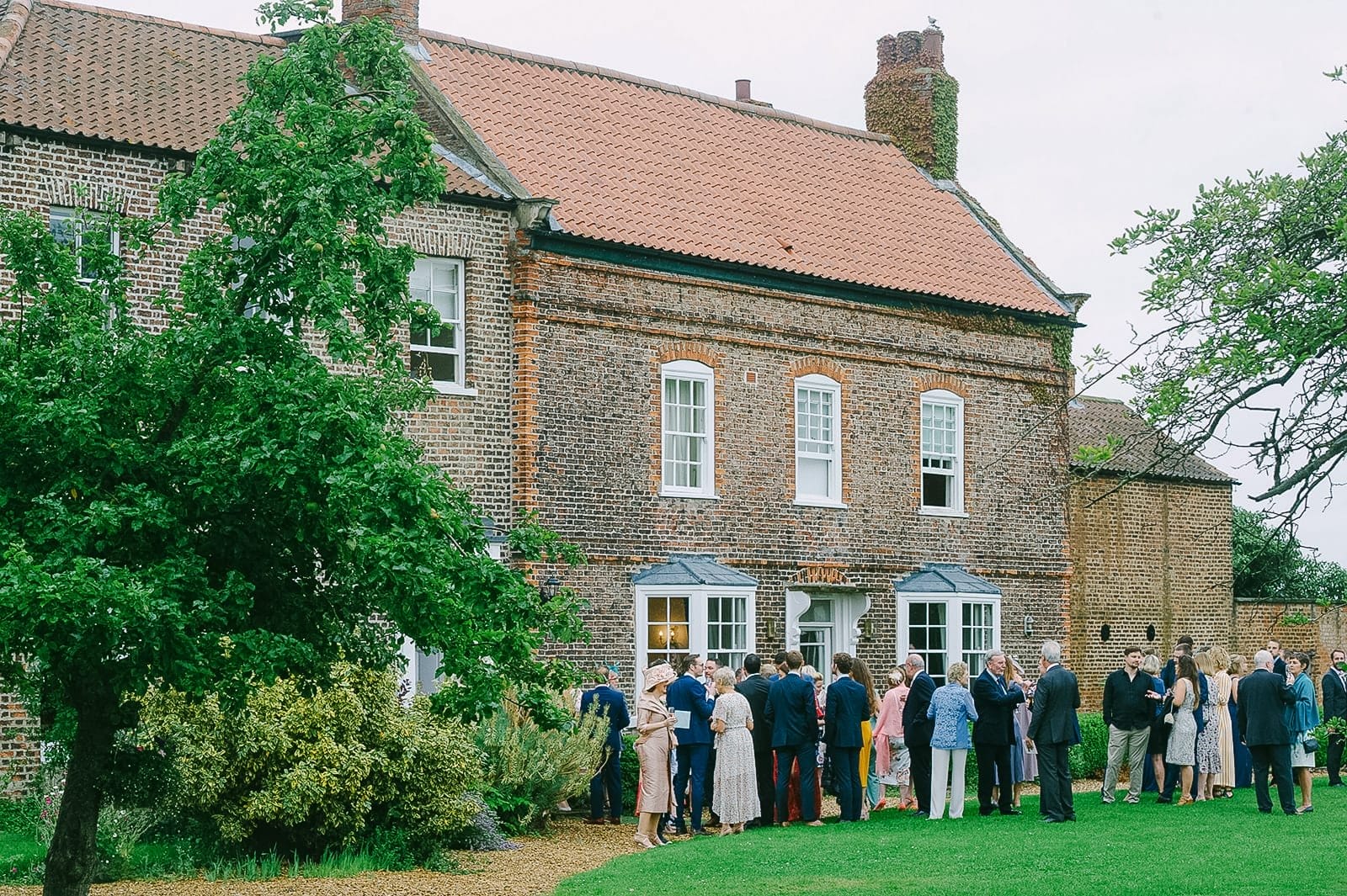 A group of people dressed in formal attire gather on a lawn outside Hornington Manor, a large brick house with bay windows and a red-tiled roof. Lush greenery surrounds the scene, creating the perfect backdrop for a wedding photographer capturing this warm and social outdoor event.