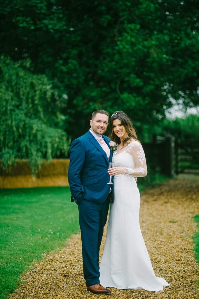 The couple beams on a gravel path at Hornington Manor, encircled by lush greenery. She dazzles in a white wedding dress while he complements her in a blue suit, their smiles bright under the sheltering trees—captured perfectly by the wedding photographer.