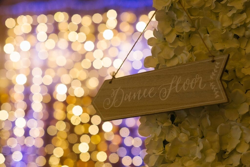 A wooden arrow sign with Dance Floor written on it hangs against a backdrop of yellow and purple bokeh lights and white flowers, capturing the elegant and festive atmosphere through the lens of a Hornington-manor-wedding-photographer.