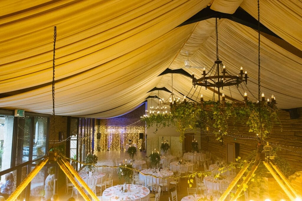 Nestled in Hornington Manor, this warmly lit wedding venue boasts a draped ceiling and chandeliers. Round tables adorned with white linens and floral centerpieces grace the space, ready for an event. String lights gently illuminate the scene, perfect for capturing those special moments by a skilled photographer.