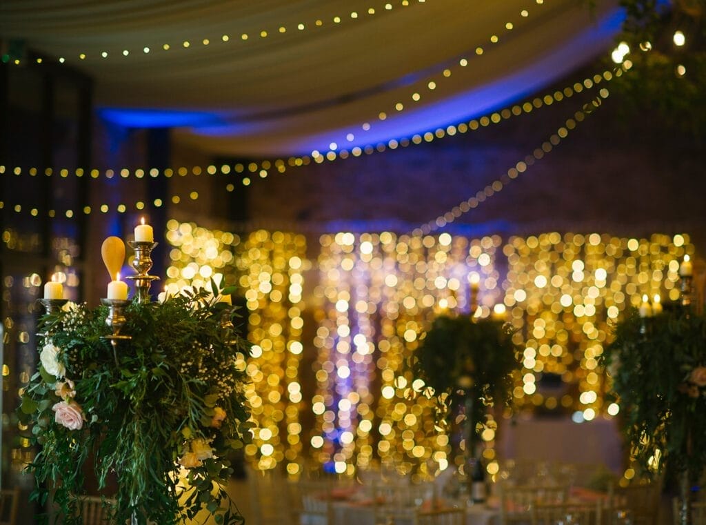 A beautifully decorated wedding venue with warm fairy lights draped across the ceiling and walls. Tall floral centerpieces with candles adorn the tables, creating an elegant and romantic atmosphere captured perfectly by a Hornington-manor-wedding-photographer.