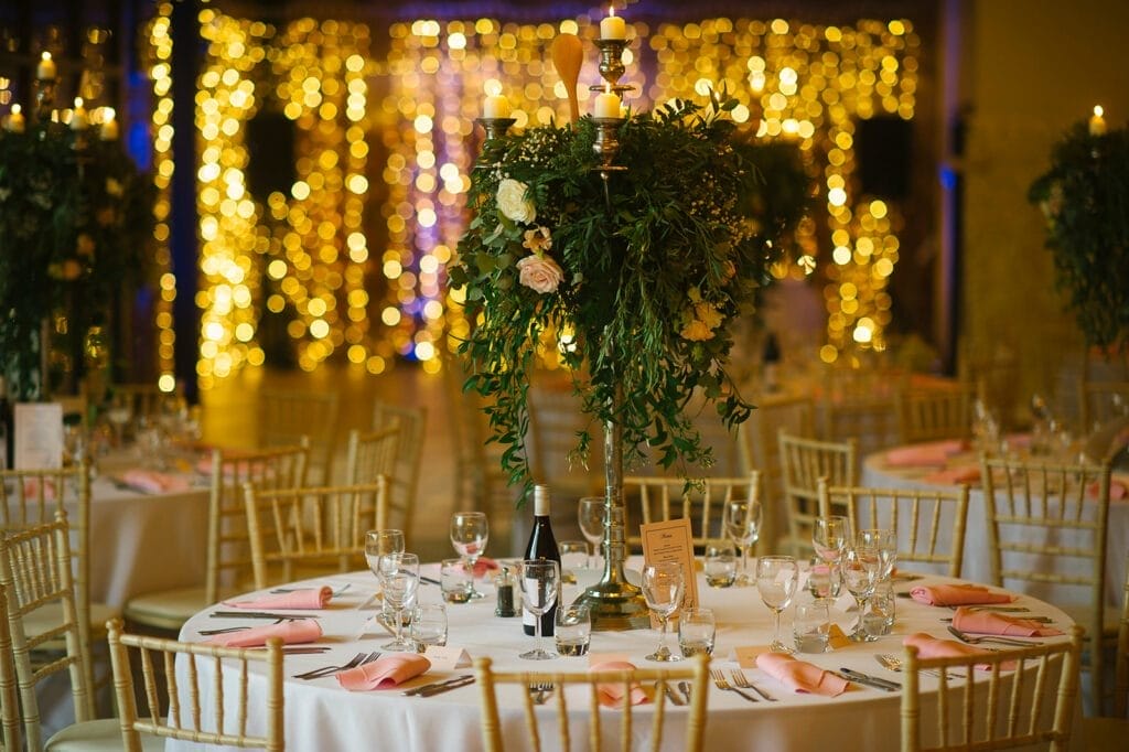 Elegant wedding reception with round tables adorned with white linens and pink napkins captured by a Hornington Manor wedding photographer. A tall floral centerpiece with greenery and candles decorates the table, while warm string lights create a cozy ambiance in the background.