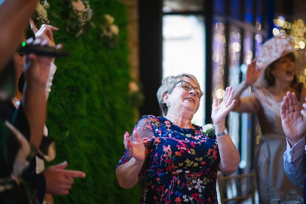 At a Hornington Manor wedding, a woman in a floral dress joyfully dances with her hands raised at the lively gathering. Others join in the celebration, amid festive decor and plants, perfectly captured by the wedding photographer.