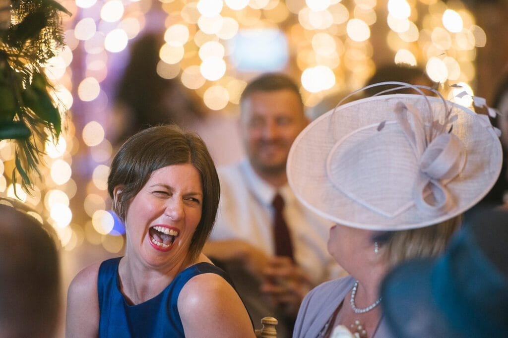 A woman in a blue dress laughs joyfully while chatting with another woman in a hat at an event. The background, blurred with warm, twinkling lights, hints at elegance. A smiling man in a tie stands distantly—an ideal scene for a Hornington-manor-wedding-photographer to capture.