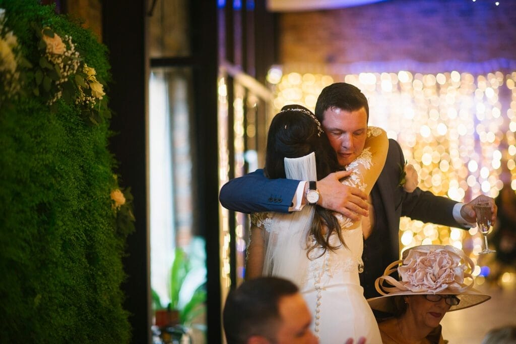 At Hornington Manor, a bride and groom embrace warmly during their wedding reception. The backdrop glows with soft string lights, while a woman in a flowered hat sits nearby amidst lush green foliage decorating the venue.