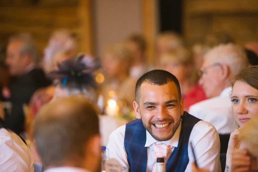 A smiling man in formal attire with a pink tie sits at a table during a lively event at Hornington Manor. He is surrounded by others engaged in conversation, all basking in the warm, softly-lit atmosphere captured by the wedding photographer.