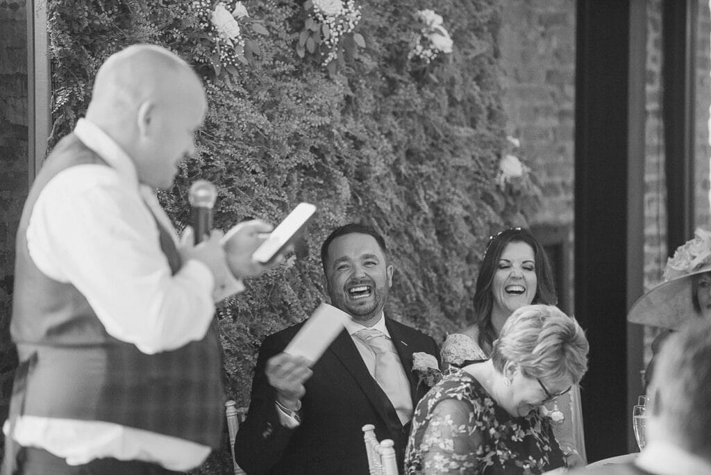 A man in a suit and a woman sit at a table, both laughing during a speech at Hornington Manor. Another man stands with a microphone and paper, speaking to an audience. A wall with greenery enhances the background. The black-and-white image captures the elegance of wedding photography.