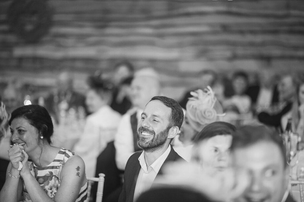 A black and white photo captures a smiling man in a suit at what appears to be a Hornington Manor wedding. Surrounded by guests focused on something off-camera, the background is elegantly blurred with tables lending an air of sophistication.