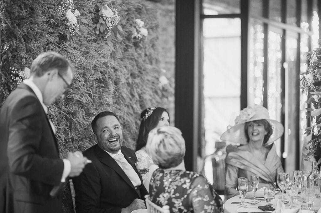 A black-and-white photo captures the charm of a Hornington Manor wedding reception. A man stands, speaking to seated guests who are laughing. A woman in a large hat sits nearby, smiling. The table is set with glasses and floral decor adorns the wall behind them.