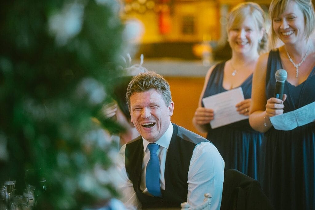 A man in a suit and tie sits laughing at a gathering, effortlessly captured by the Hornington Manor wedding photographer. Two women in blue dresses stand nearby—one with a microphone, the other holding a paper—as the softly blurred background enhances the festive atmosphere.