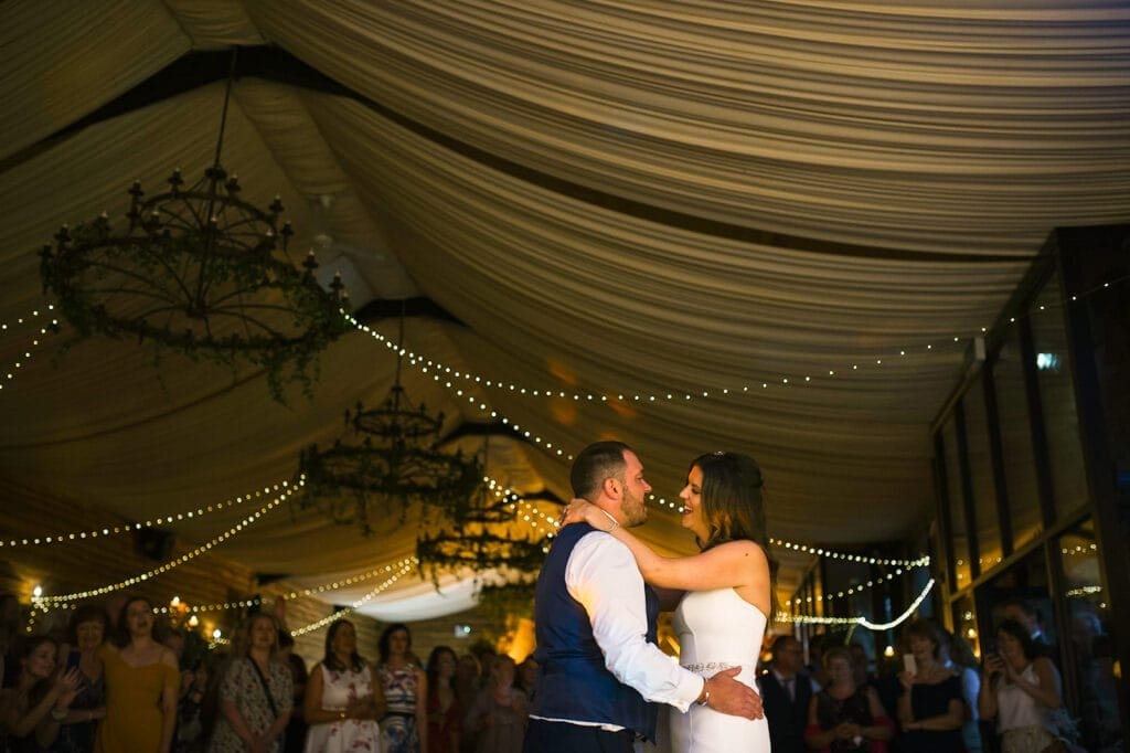 A couple dances gracefully under a draped tent adorned with string lights and chandeliers at Hornington Manor. The bride’s white dress glows, complementing the grooms blue vest and white shirt, as guests watch against a softly lit background. This scene is perfectly captured by the wedding photographer.