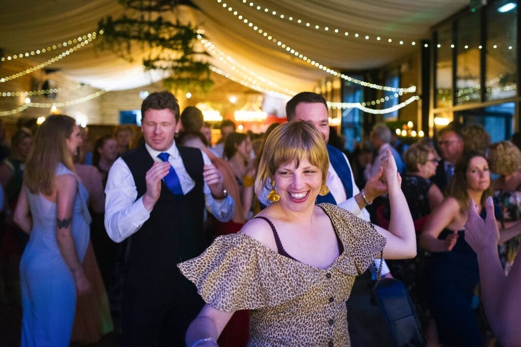 A lively indoor party scene captured by a skilled Hornington Manor wedding photographer features people dancing under string lights. A smiling woman in a leopard print dress stands in the foreground, while others, including a man in a suit and tie, enjoy the festive atmosphere of celebration.