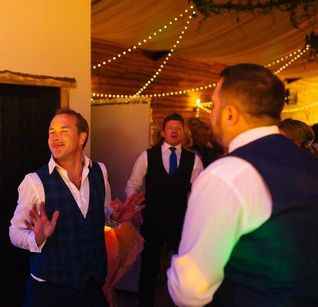 At Hornington Manor, a wedding photographer captures the joy of people dancing amid twinkling string lights. One person in a white shirt and blue vest sways with closed eyes, fully immersed in the moment, while others, donned in formal attire, share smiles and laughter.