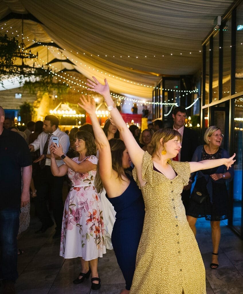 A group of people dancing joyfully at a festive Hornington Manor wedding event with warm lights and decorations. One woman in the foreground wearing a yellow dress has her arms raised. Others behind her are also dancing or taking photos, captured perfectly by the wedding photographer amid smiles and cheerful expressions.