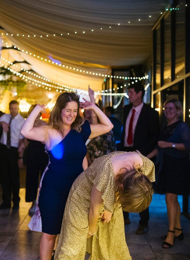 Two women are dancing joyfully at a vibrant Hornington Manor wedding under string lights. One, in a navy dress, raises her arms while the other, in a patterned dress, leans forward laughing. The lively atmosphere has everyone in the background enjoying the celebration to its fullest.