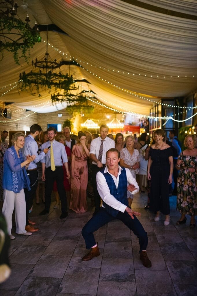 A man in a vest and tie dances energetically on the dance floor at Hornington Manor. Guests in formal attire surround him, smiling and clapping. The venue, captured beautifully by the wedding photographer, is adorned with string lights and draped fabric.