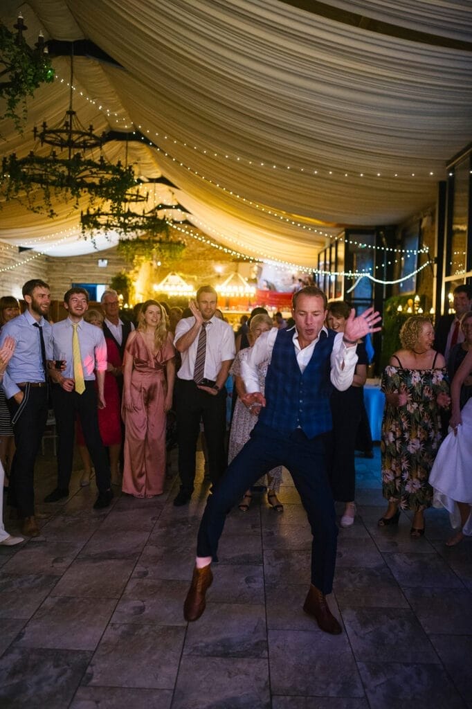 A man energetically dances in the center of a crowd at a lively indoor event with string lights and a draped ceiling. As guests in formal attire smile and watch, the scene is perfectly captured by the Hornington Manor wedding photographer.