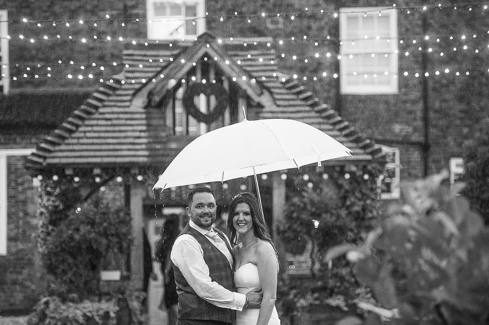 A bride and groom stand under a white umbrella, smiling at the camera. Behind them, Hornington Manors brick facade glows with string lights, creating an enchanting evening scene captured perfectly by their wedding photographer.