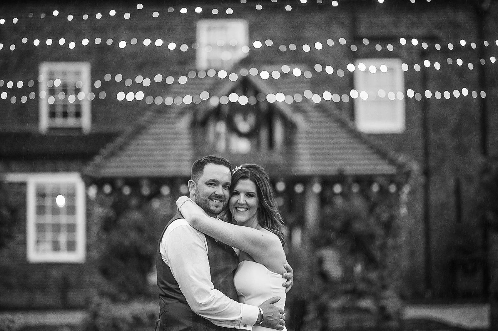 A couple embraces and smiles under string lights in front of a brick building, captured by a Hornington Manor wedding photographer. The black-and-white scene with blurred lights creates a romantic atmosphere.