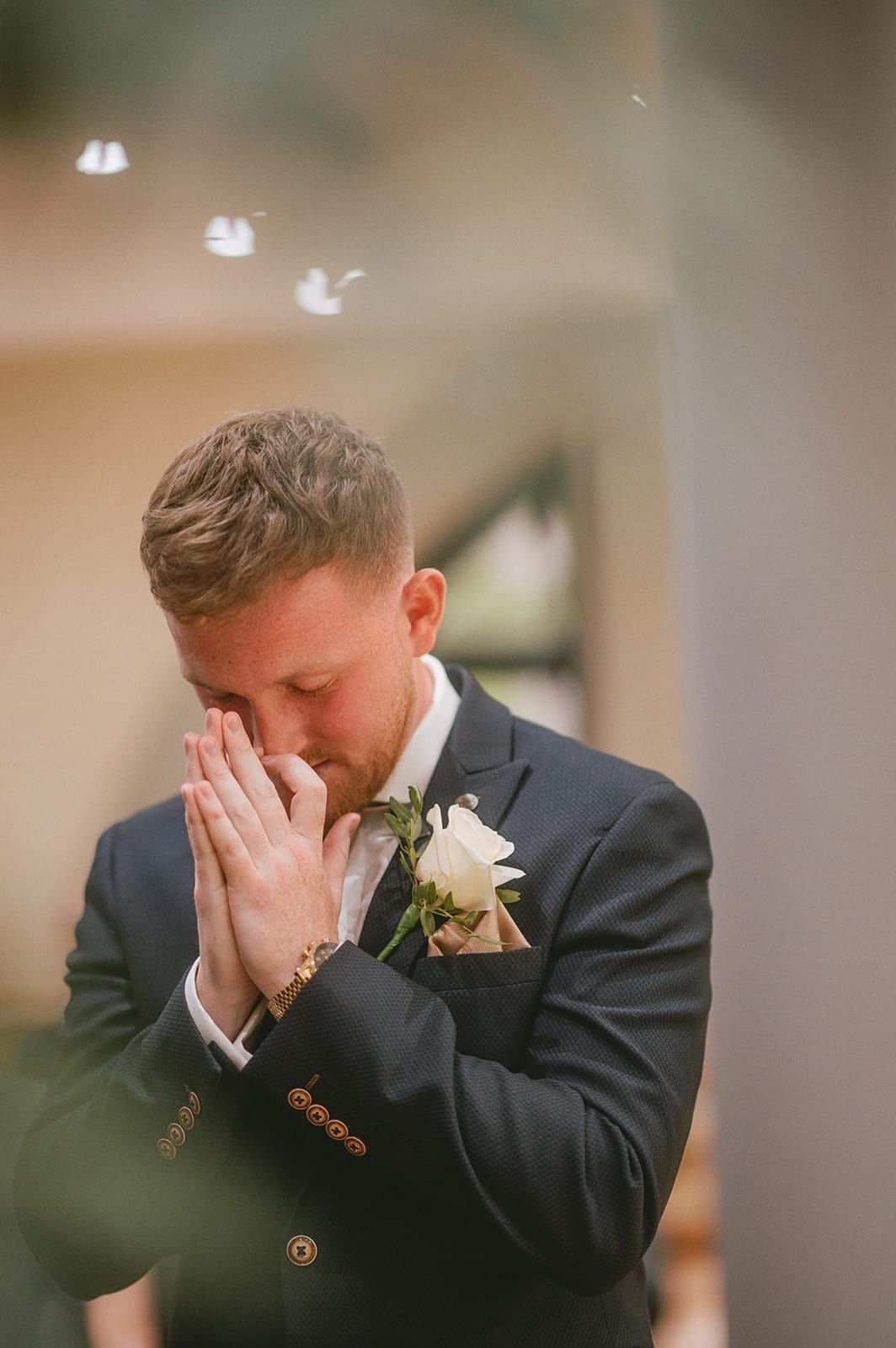 A groom in a suit stands with hands pressed together near his face, eyes closed in a contemplative moment. A white rose boutonniere adorns his jacket. Captured by the wedding photographer, the softly blurred background focuses attention on his expression.